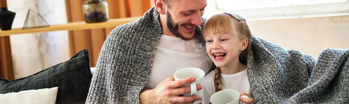 Eine Vater und seine Tochter sitzen in eine Decke gekuschelt auf dem Sofa und trinken etwas aus Tassen