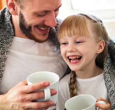 Eine Vater und seine Tochter sitzen in eine Decke gekuschelt auf dem Sofa und trinken etwas aus Tassen