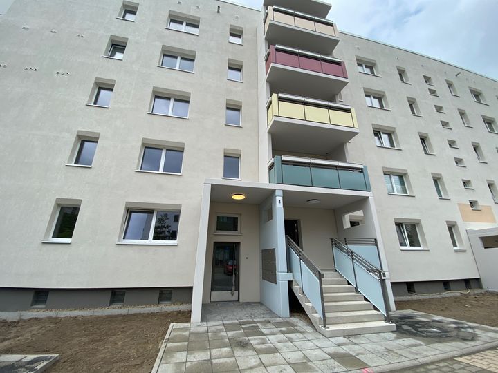 Modernisierter Eingangsbereich eines sanierten Plattenbaus im Hertha-Thiele-Weg mit neu gepflasterten Boden, weißer Fassade und Balkonen.