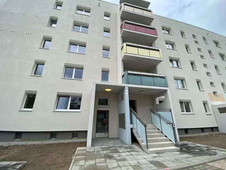 Modernisierter Eingangsbereich eines sanierten Plattenbaus im Hertha-Thiele-Weg mit neu gepflasterten Boden, weißer Fassade und Balkonen.