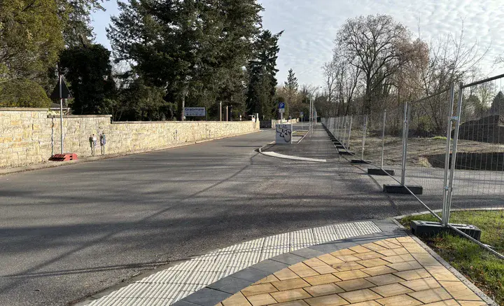 Straßenkurve mit taktilem Leitsystem am Gehweg, links eine Steinmauer und rechts ein eingezäuntes Gelände mit Bäumen.