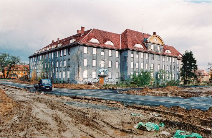 Ein altes, zerfallenes Kasernen-Gebäude. Davor eine schmale Straße und viel aufgewühlte Erde.