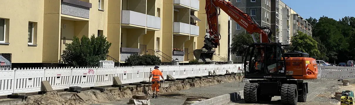 Aufgerissene Straße und Baustellenfahrzeuge vor einem Mietshaus.