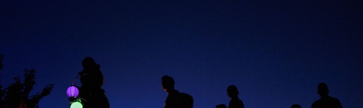 Menschen mit einer Laterne gehen in einer Reihe. Im Hintergrund ist dunkler Nachthimmel zu sehen. 
