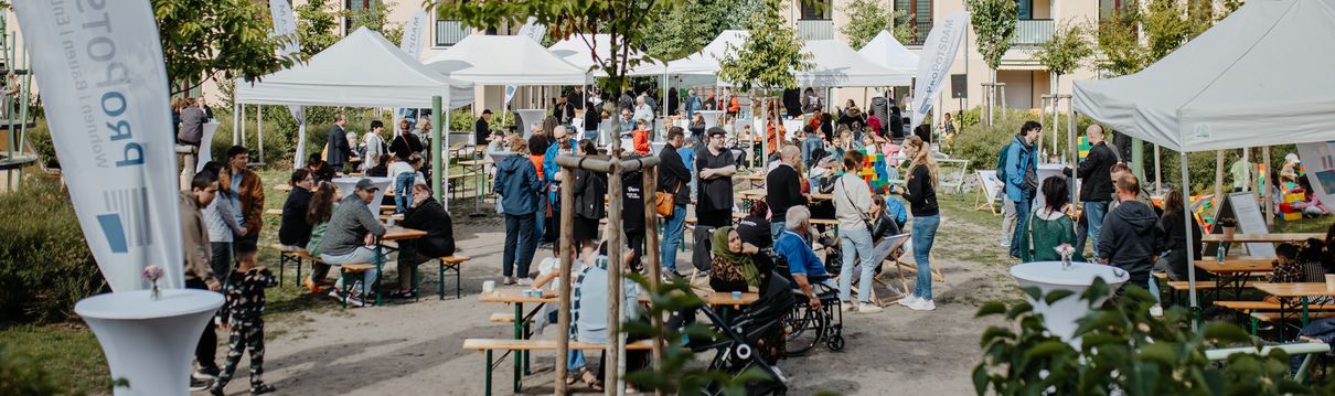 Belebtes Nachbarschaftsfest im begrünten Innenhof einer modernen Wohnanlage mit weißen Pavillons, Bierbänken und vielen Gästen