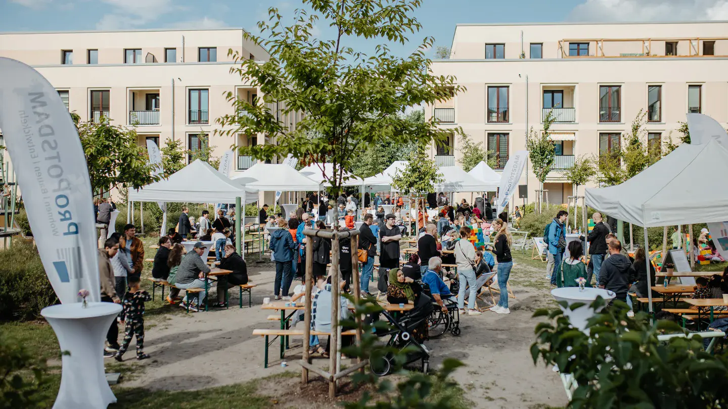 Belebtes Nachbarschaftsfest im begrünten Innenhof einer modernen Wohnanlage mit weißen Pavillons, Bierbänken und vielen Gästen