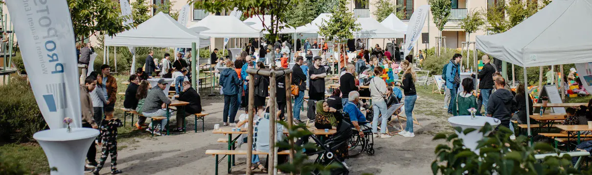 Belebtes Nachbarschaftsfest im begrünten Innenhof einer modernen Wohnanlage mit weißen Pavillons, Bierbänken und vielen Gästen