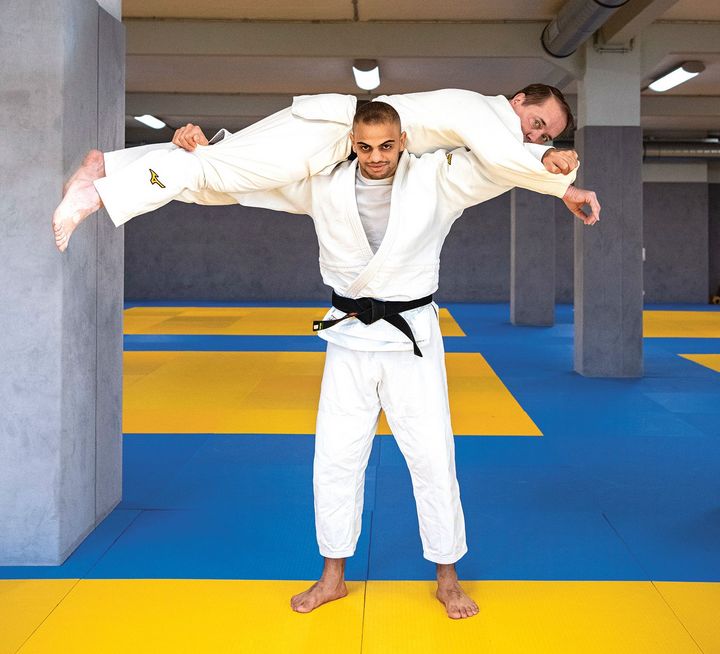 Zwei Judo-Kämpfer im weißen Anzug auf gelb-blauen Trainingsmatten in einer Sporthalle.