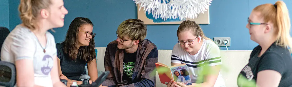 Blick auf eine Sitzecke vor blauer Wand: Ein Mädchen im Rollstuhl, zwei Mädchen und ein Junge auf einem Sofa und ein Mädchen in einem Sessel sitzen zusammen, unterhalten sich, lächeln, ein Mädchen mit Brille liest