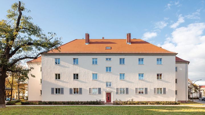 Helle Außenansicht eines sanierten Wohnkomplexes mit roter Eingangstür, Fensterläden in der untersten Etage und vielen kleinen, symmetrisch angeordneten Fenstern bis zum orangeroten Dach