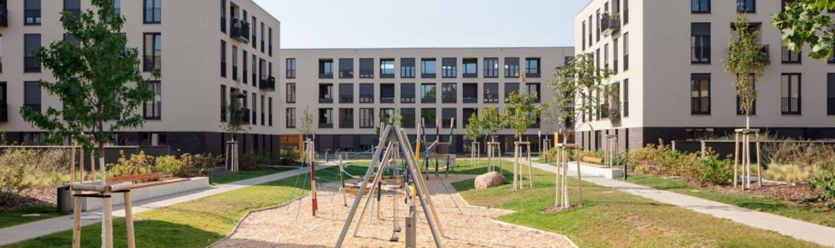 Ein Spielplatz mit Sand und Spiel- und Klettergeräten und drei mehrstöckige Wohnhäuser im Hintergrund.
