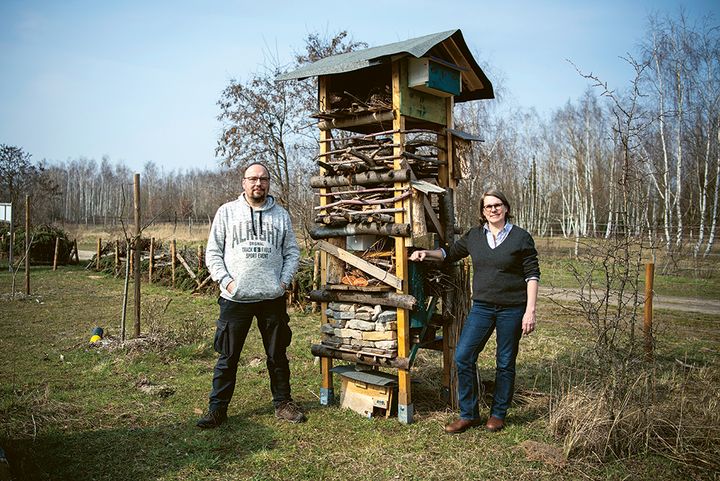 Ein Mann und eine Frau stehen in einer grünen Umgebung, zwischen ihnen steht ein großes Insektenhotel