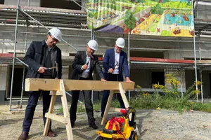 Vertreter der ProPotsdam und der Oberbürgermeister der Stadt stehen vor einem neu errichteten Gebäude und schlagen Nägel in einen Holzbalken ein. 