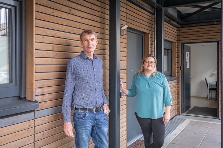 Ein Mann und eine Frau stehen vor einer mit Holz verkleideten Außenwand und blicken in die Kamera.