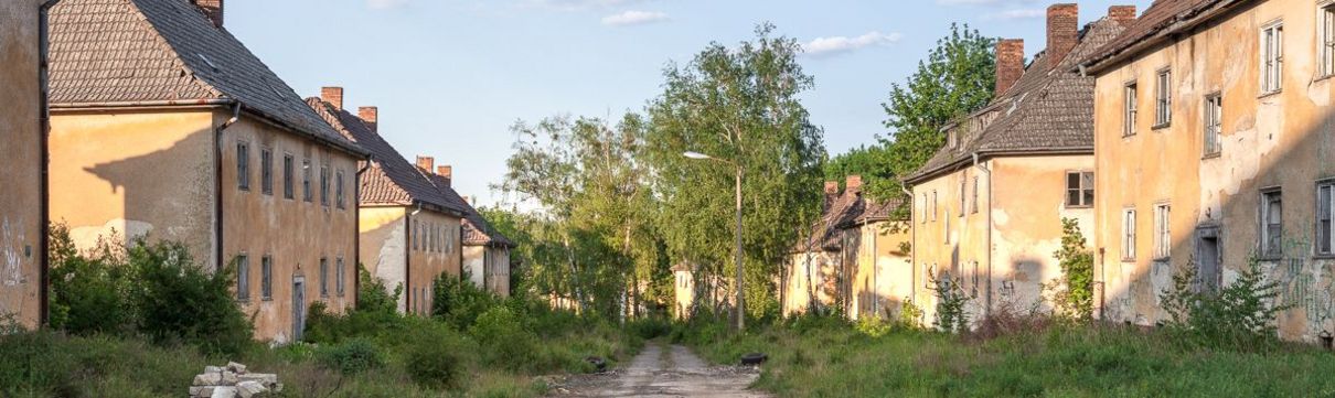 Zu sehen ist eine unbefestigte Straße und links und rechts davon verfallene Häuser. Im Hintergrund stehen Birken vor einem blauen Himmel.