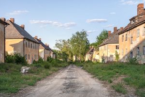 Zu sehen ist eine unbefestigte Straße und links und rechts davon verfallene Häuser. Im Hintergrund stehen Birken vor einem blauen Himmel.