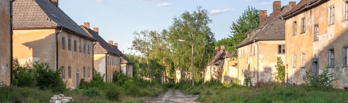 Zu sehen ist eine unbefestigte Straße und links und rechts davon verfallene Häuser. Im Hintergrund stehen Birken vor einem blauen Himmel.