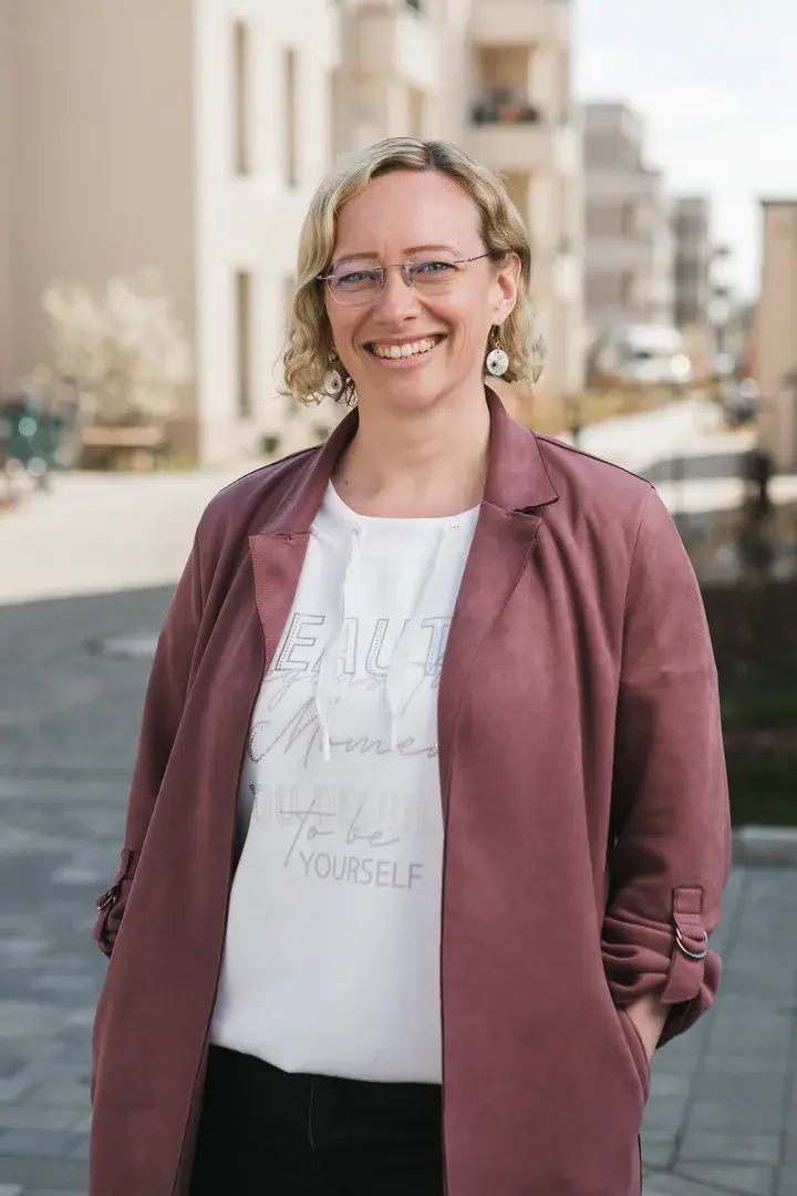 Cindy Plateo Auf dem Foto sieht man eine blonde Frau mit Brille, die lächelt. Im Hintegrund stehen Häuser.