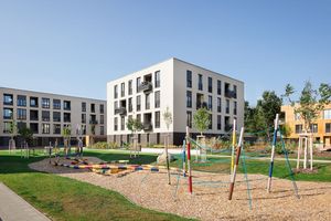 Ein Kinderspielplatz mit Klettergerüsten und anderen Spielgeräten. Im Hintergrund sind Wohnhäuser zu sehen. 