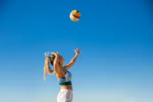 Auf dem Bild ist eine Frau im Sportoutfit zu sehen, die vor einem blauen Himmel zum Schlag auf einen Volleyball ausholt.