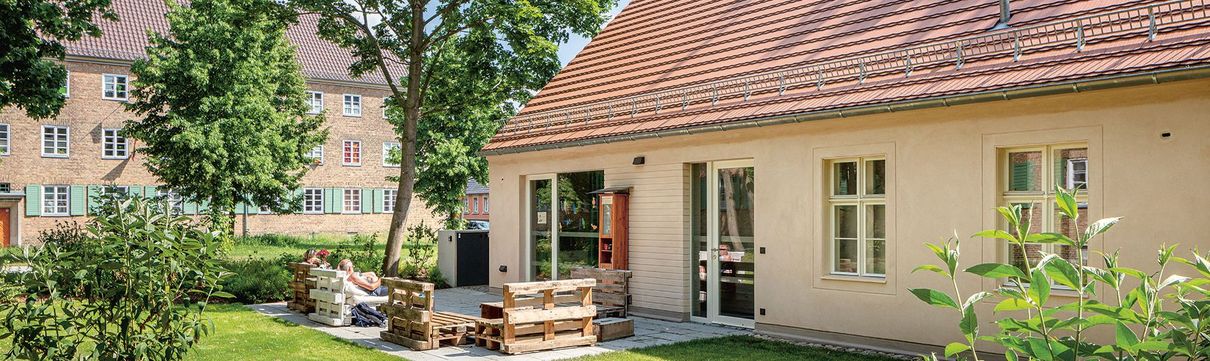 Rückansicht eines hellen Häuschens mit Blick auf die Terrasse, die mit Palettenmöbeln ausgestattet zwei Personen zum Verweilen einlädt