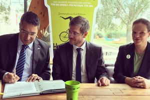 Der Geschäftsführer der ProPotsdam, der gerade etwas unterzeichnet, sitzt mit dem Oberbürgermeister und der Vorsitzenden der Bürgerstiftung Potsdam an einem Tisch. Im Hintergrund ist ein großer Kaffeebecher aus Pappe zu sehen.