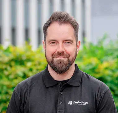Portrait einer Person im schwarzen ProPotsdam-Poloshirt vor grüner Hecke und modernem Gebäude im Hintergrund.