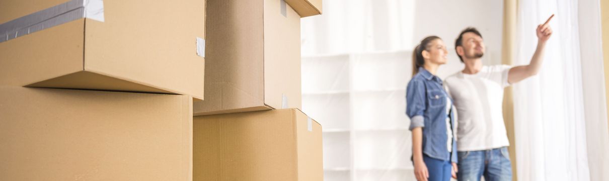 In der linken Bildhälfte stehen Stapel großer Umzugskisten im Fokus. In der rechten Bildhälfte sieht man unscharf im Hintergrund eine Frau und einen Mann Anfang 30, der der der Frau mit dem Finger etwas zeigt, dass sich draußen vor dem Fenster befindet.