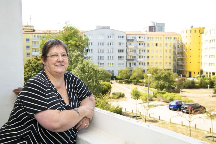 Eine Frau steht auf einem Balkon, einen Arm auf die Brüstung gestützt und lächelt in die Kamera. Im Hintergrund sind Wohngebäude zu sehen.
