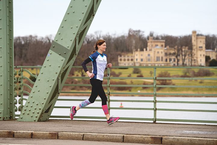 Joggerin läuft über eine Brücke mit grünen Stahlträgern in deren Hintergrund ein kleines Schloss sichtbar ist