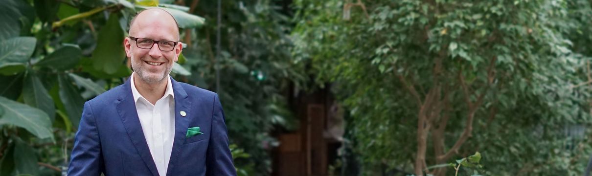 Mann in blauem Anzug und mit Brille steht lächeln in der Biosphäre Potsdam, Dschungelhintergrund