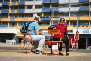 Zwei erwachsene Personen mit Kopfhörern auf den Ohren sitzen auf orangefarbenen Klappstühlen mit Blick auf einen Plattenbau, vor dem eine Tänzerin im roten Kleid performt.