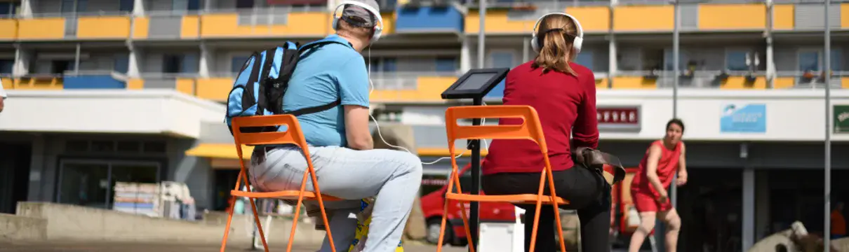 Zwei erwachsene Personen mit Kopfhörern auf den Ohren sitzen auf orangefarbenen Klappstühlen mit Blick auf einen Plattenbau, vor dem eine Tänzerin im roten Kleid performt.