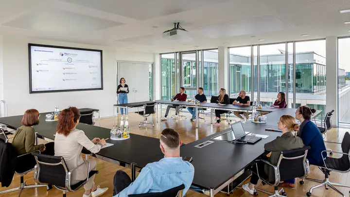 Arbeitsberatung einer größeren Personengruppe an U-förmig angeordneten Tischen in einem großen, hellen Raum