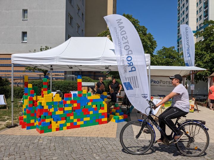 Stand mit weißem Dach in Plattenbau-Siedlung vor dem aus bunten XXL-Bausteinen ein großer Bau steht, im Vordergrund fährt ein Radfahrer